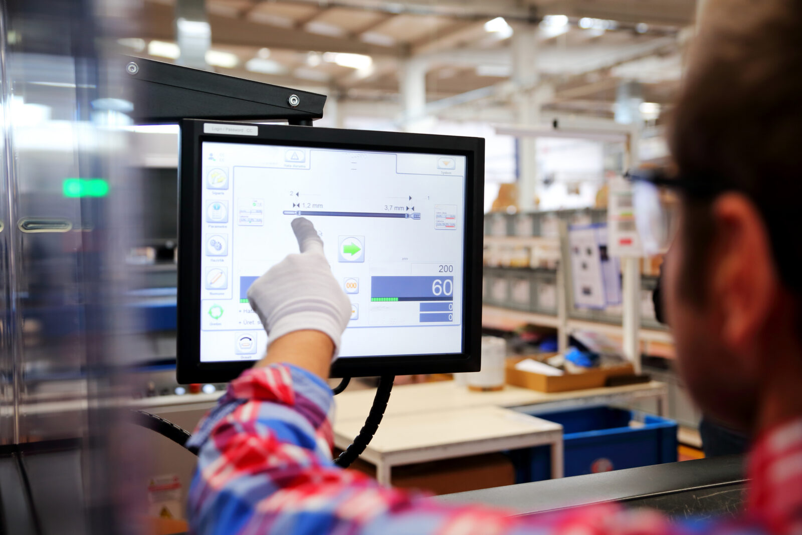 A factory worker wearing a glove and a red plaid shirt pointing at a computer touchscreen interface controlling industrial machinery.
