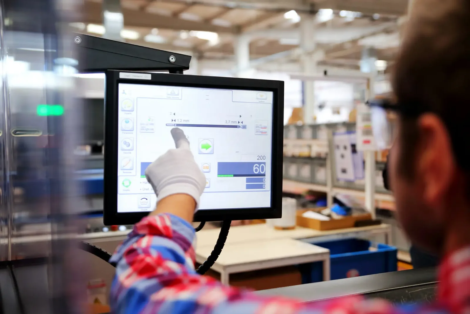 A factory worker wearing a glove and a red plaid shirt pointing at a computer touchscreen interface controlling industrial machinery.