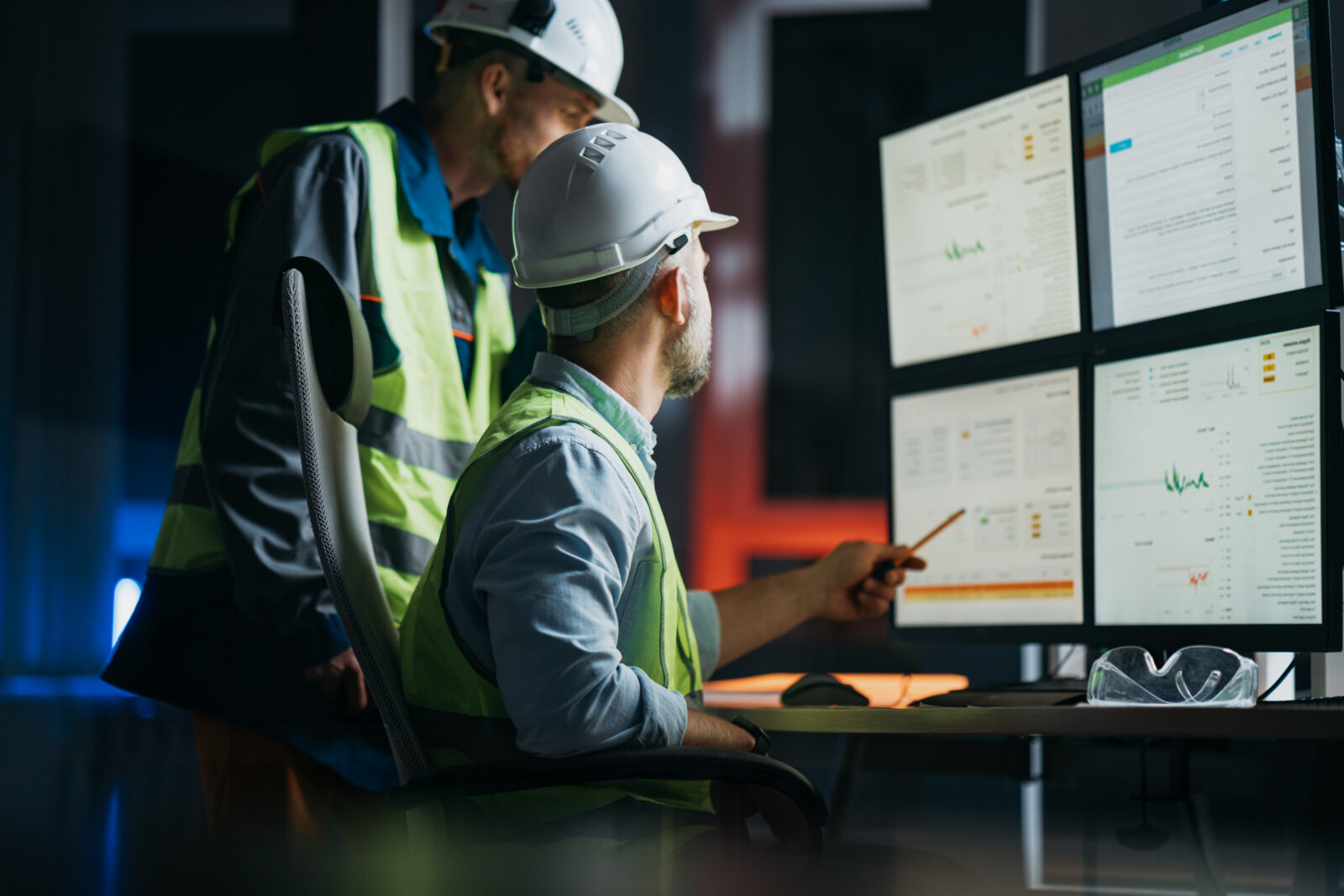 Two industrial engineers in white hard hats and neon safety vests analyzing telemetry data on a multi-monitor control desk at night.