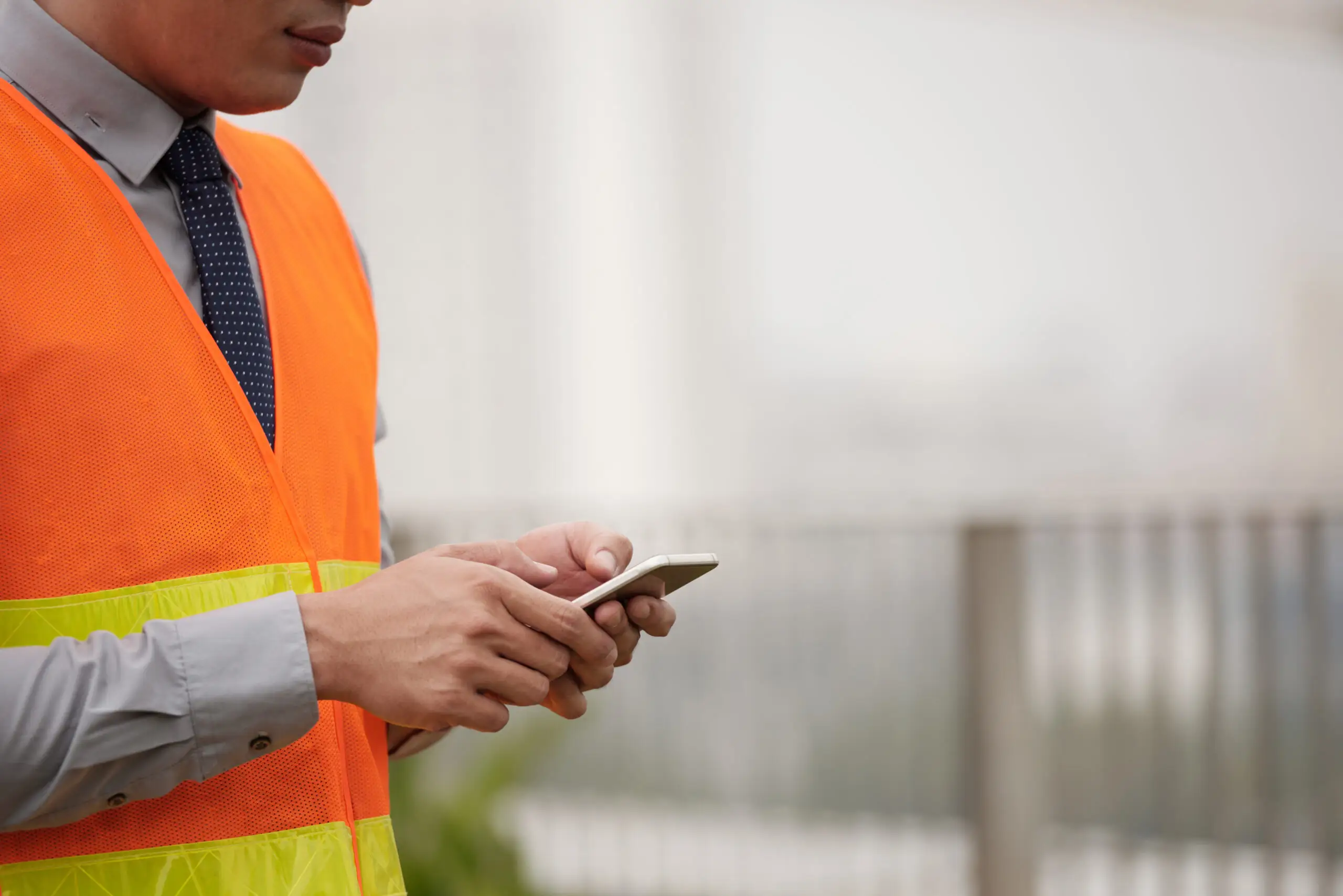 A professional in a high-visibility orange safety vest, grey dress shirt, and tie, typing on a smartphone outdoors.