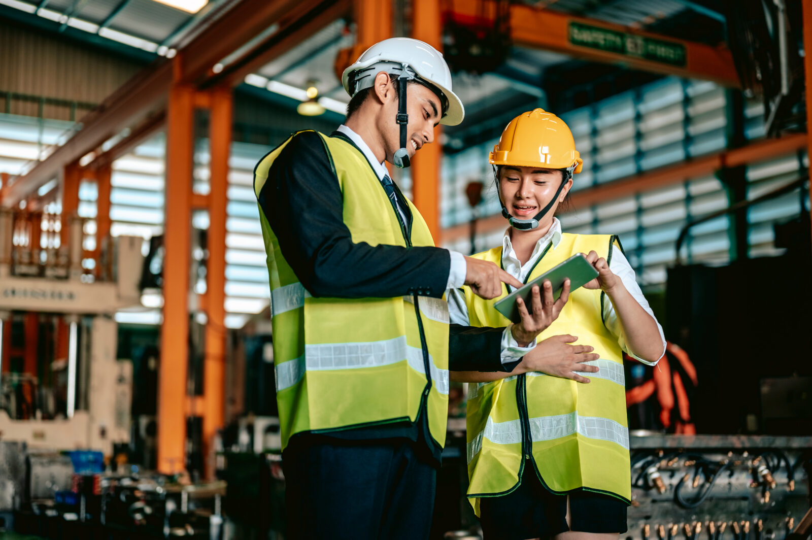 Two factory workers wearing hard hats and safety vests standing together in an industrial facility while reviewing information on a digital tablet.