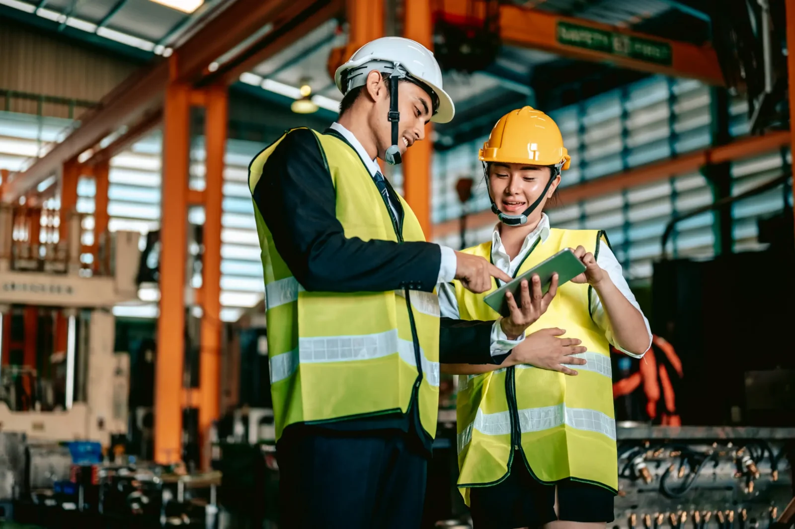Two factory workers wearing hard hats and safety vests standing together in an industrial facility while reviewing information on a digital tablet.
