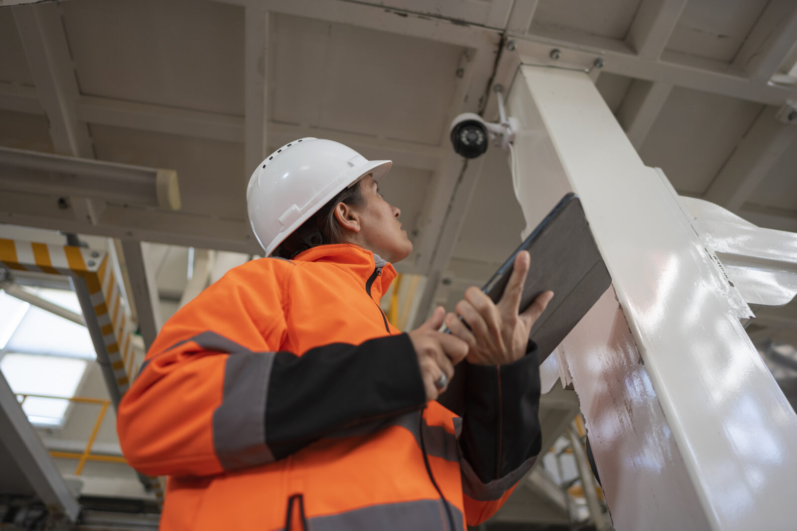 A worker wearing a white hard hat and bright orange jacket holding a tablet while inspecting a ceiling-mounted security camera inside a facility.