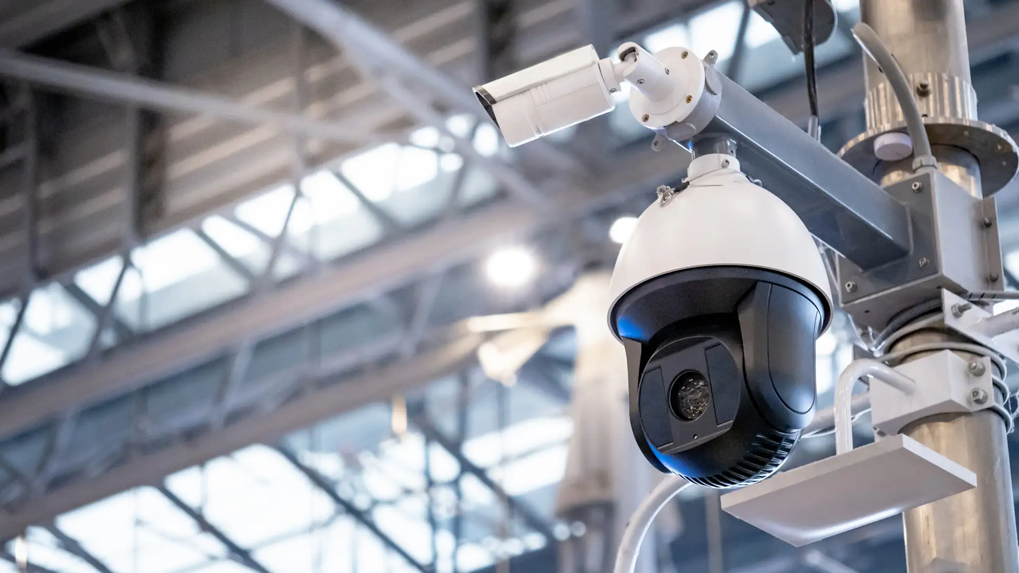 A close-up of a security dome camera and a bullet camera mounted on a metal pole inside a high-ceilinged building.