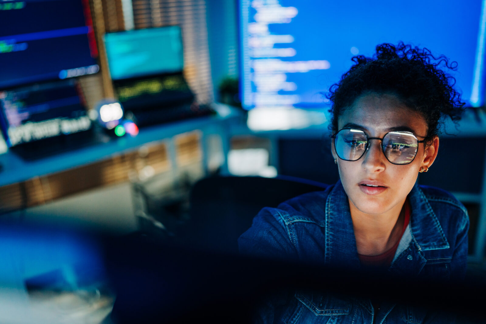 A female software developer wearing glasses looking intently at a computer screen displaying computer code in a dimly lit office.