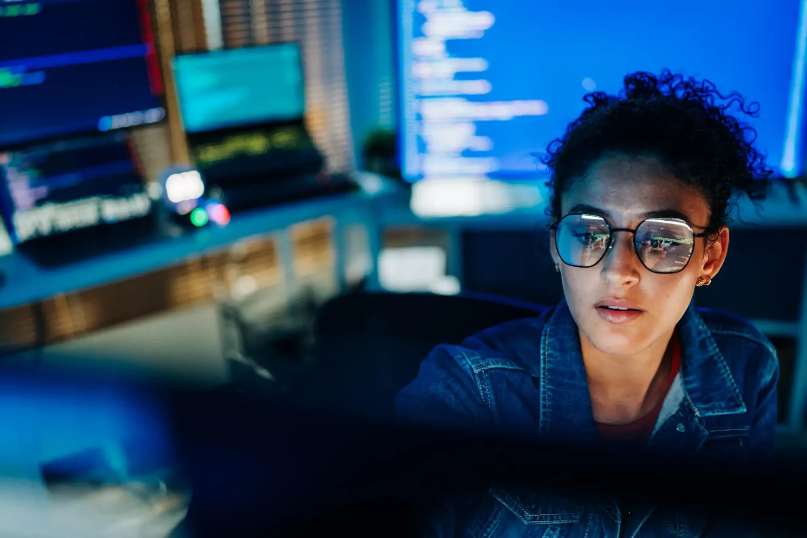 A female software developer wearing glasses looking intently at a computer screen displaying computer code in a dimly lit office.