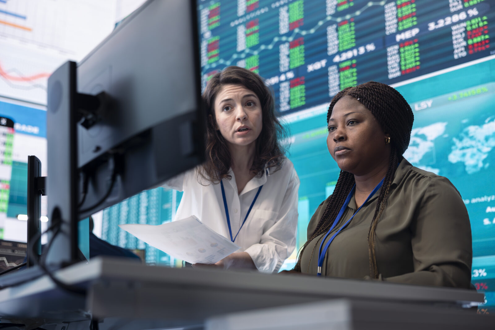Two financial analysts reviewing computer data together in a stock trading room with large screens displaying real-time market tickers.