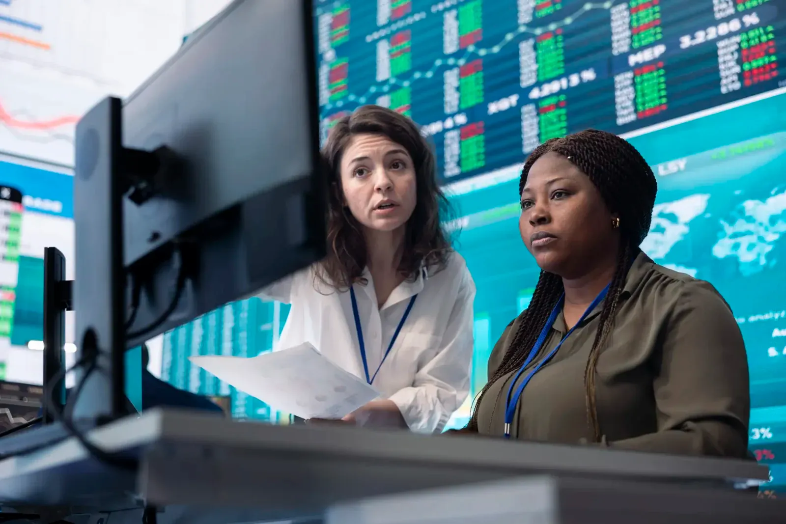 Two financial analysts reviewing computer data together in a stock trading room with large screens displaying real-time market tickers.