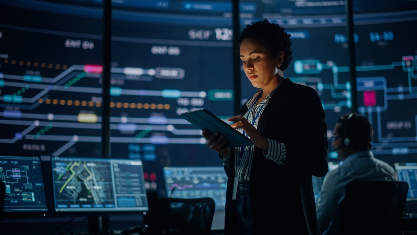 A female engineer using a tablet in a high-tech control room with large screens displaying live data maps and network schematics.