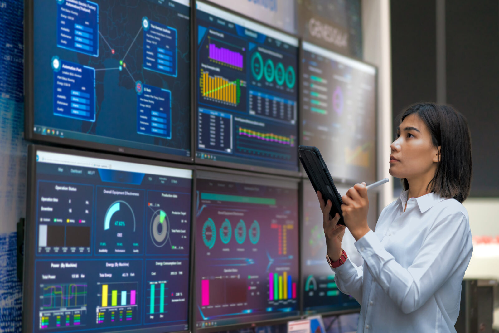 An engineer using a stylus on a tablet while looking up at a wall of monitors displaying analytics dashboards and system metrics.