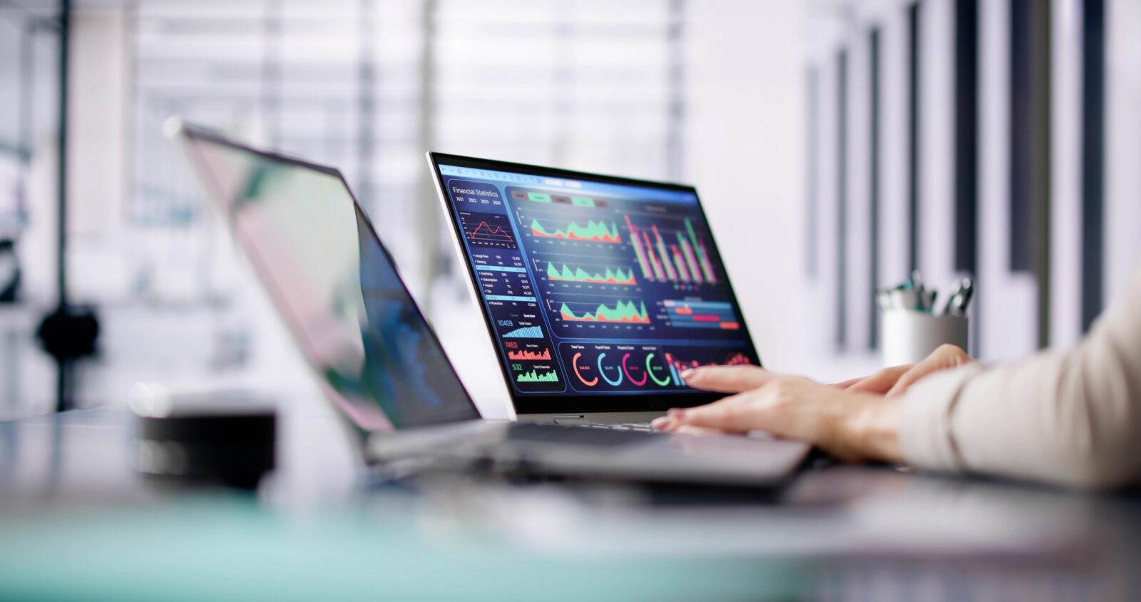 A person's hands typing on a laptop displaying colorful data charts and graphs on the screen at a modern office desk.