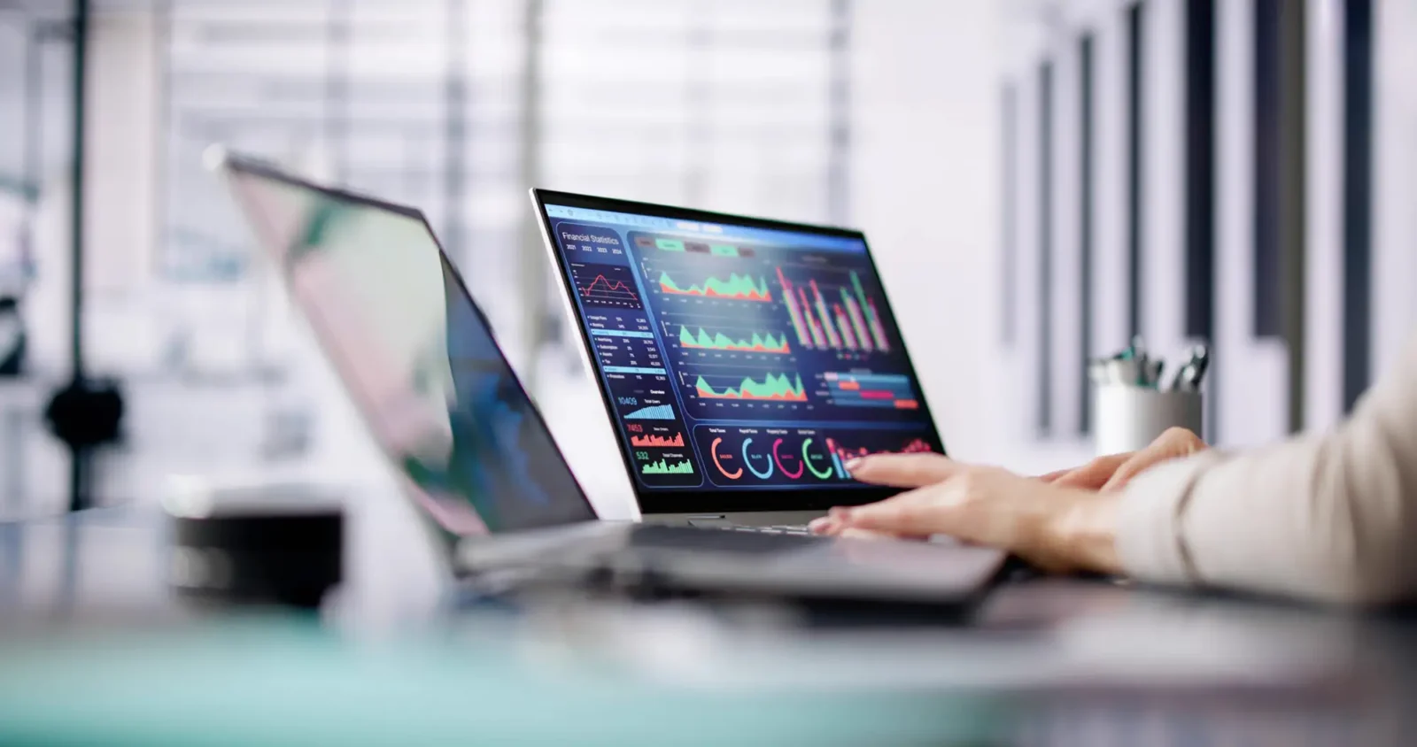 A person's hands typing on a laptop displaying colorful data charts and graphs on the screen at a modern office desk.