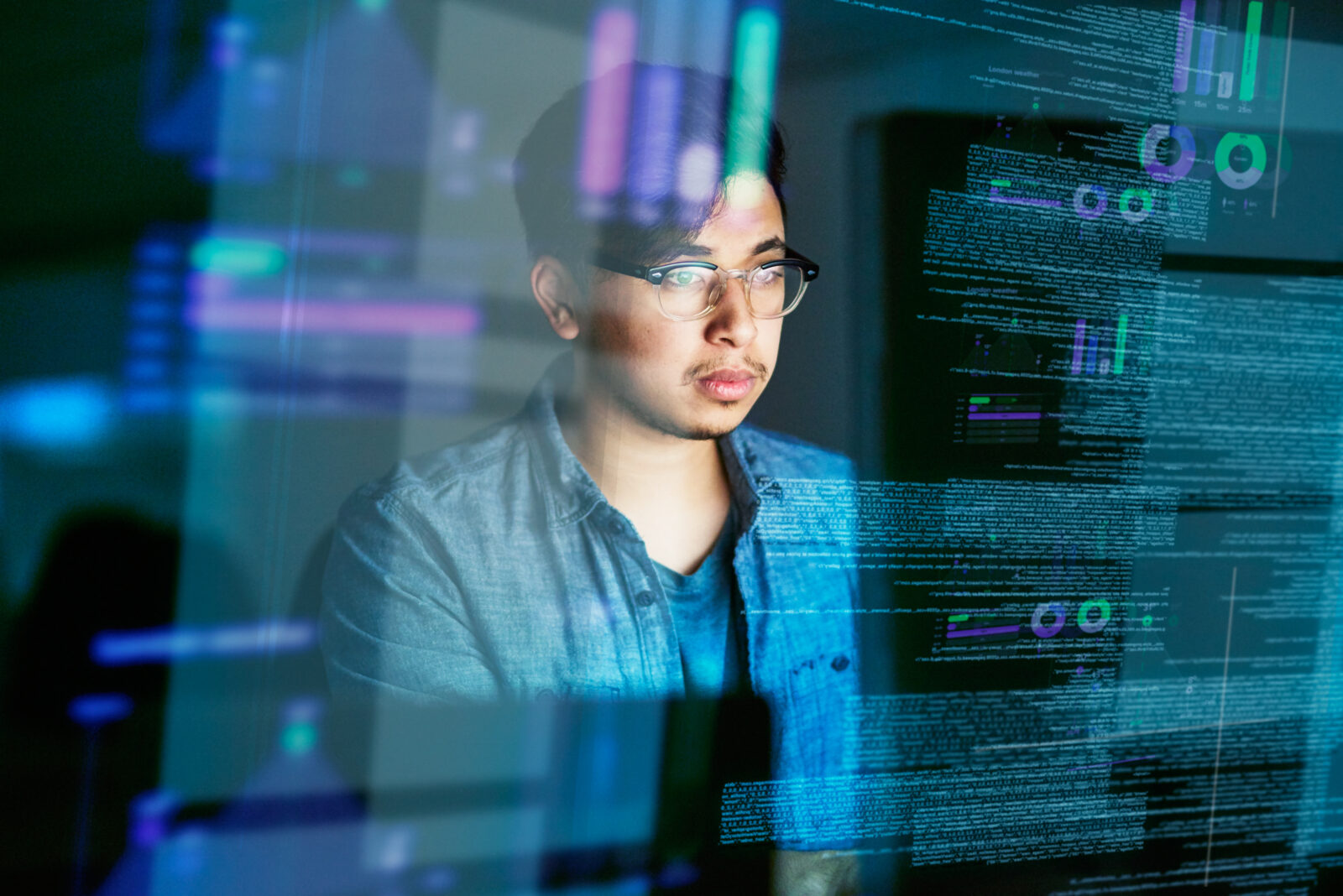 A person wearing glasses and a denim shirt looking at a screen with overlay reflections of software code, data charts, and analytics.