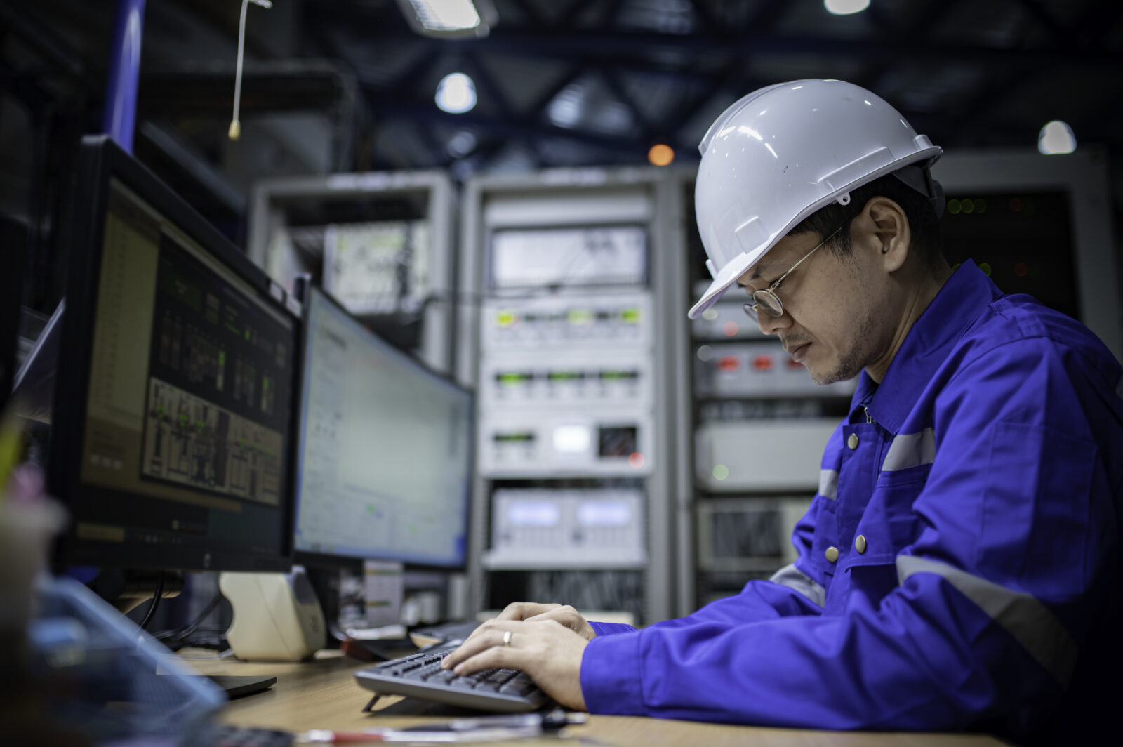 A technician wearing a white hard hat and blue work shirt typing on a computer keyboard in an industrial control room.