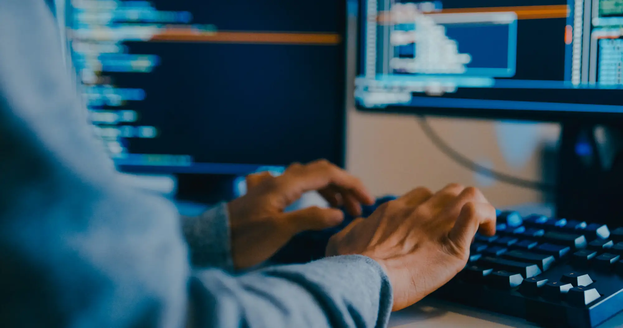 A close-up of a person's hands typing on a mechanical keyboard in front of dual computer monitors displaying lines of code.