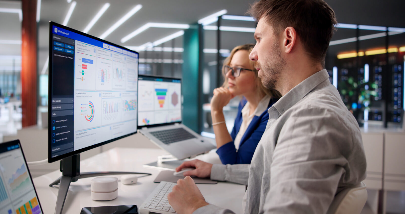 Two colleagues working together at a desk reviewing a KPI analytics dashboard on a large monitor in a modern office.