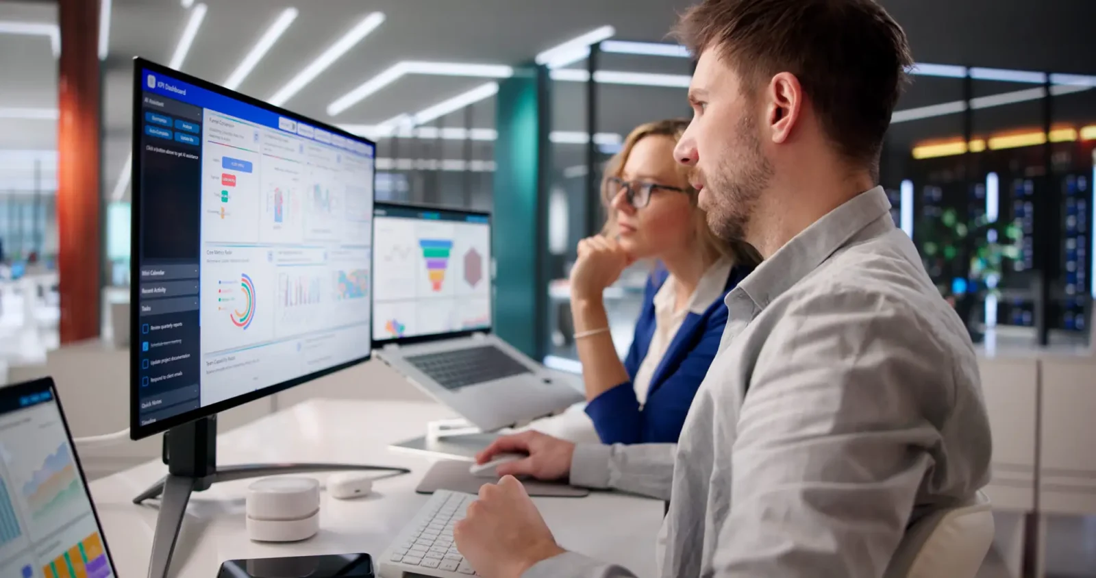 Two colleagues working together at a desk reviewing a KPI analytics dashboard on a large monitor in a modern office.