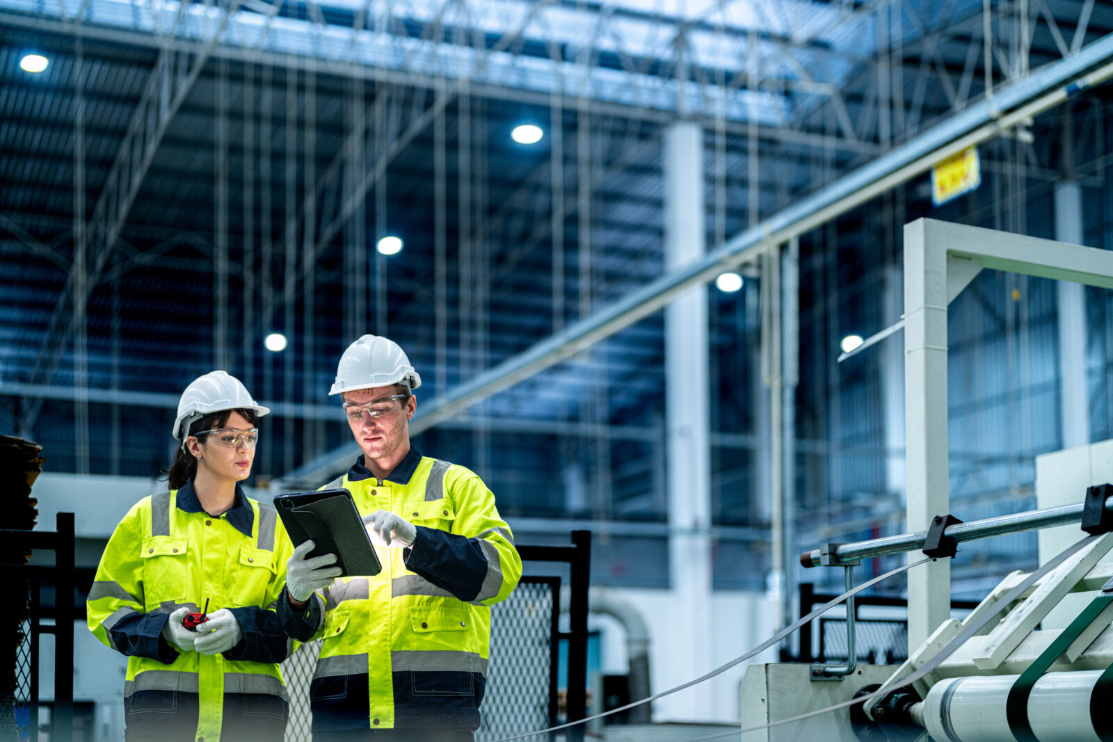 Two factory technicians in hard hats and high-visibility jackets reviewing data on a tablet inside a large manufacturing facility.