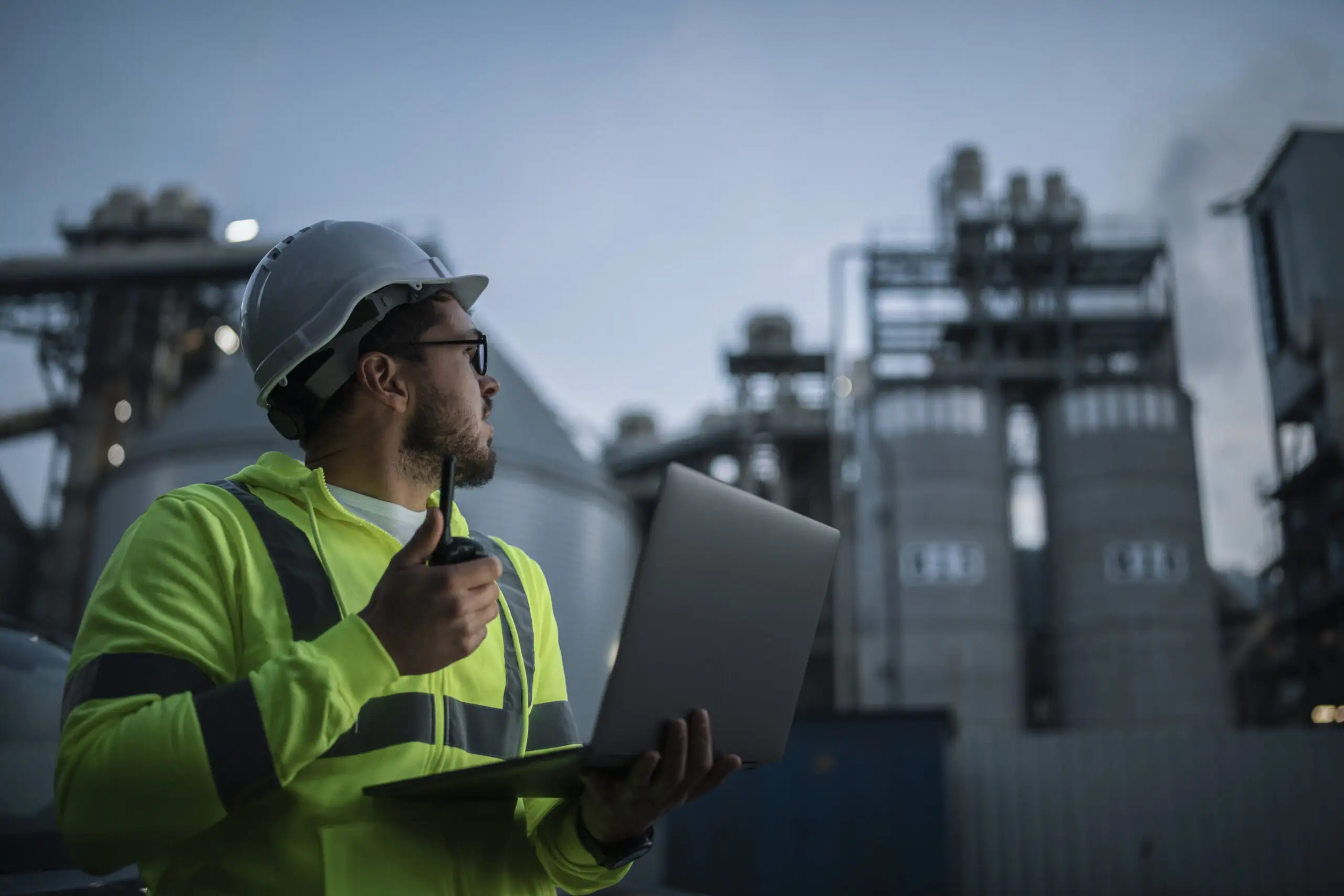 An engineer wearing a white hard hat and high-visibility jacket speaking into a walkie-talkie while holding a laptop in front of an industrial plant at dusk.