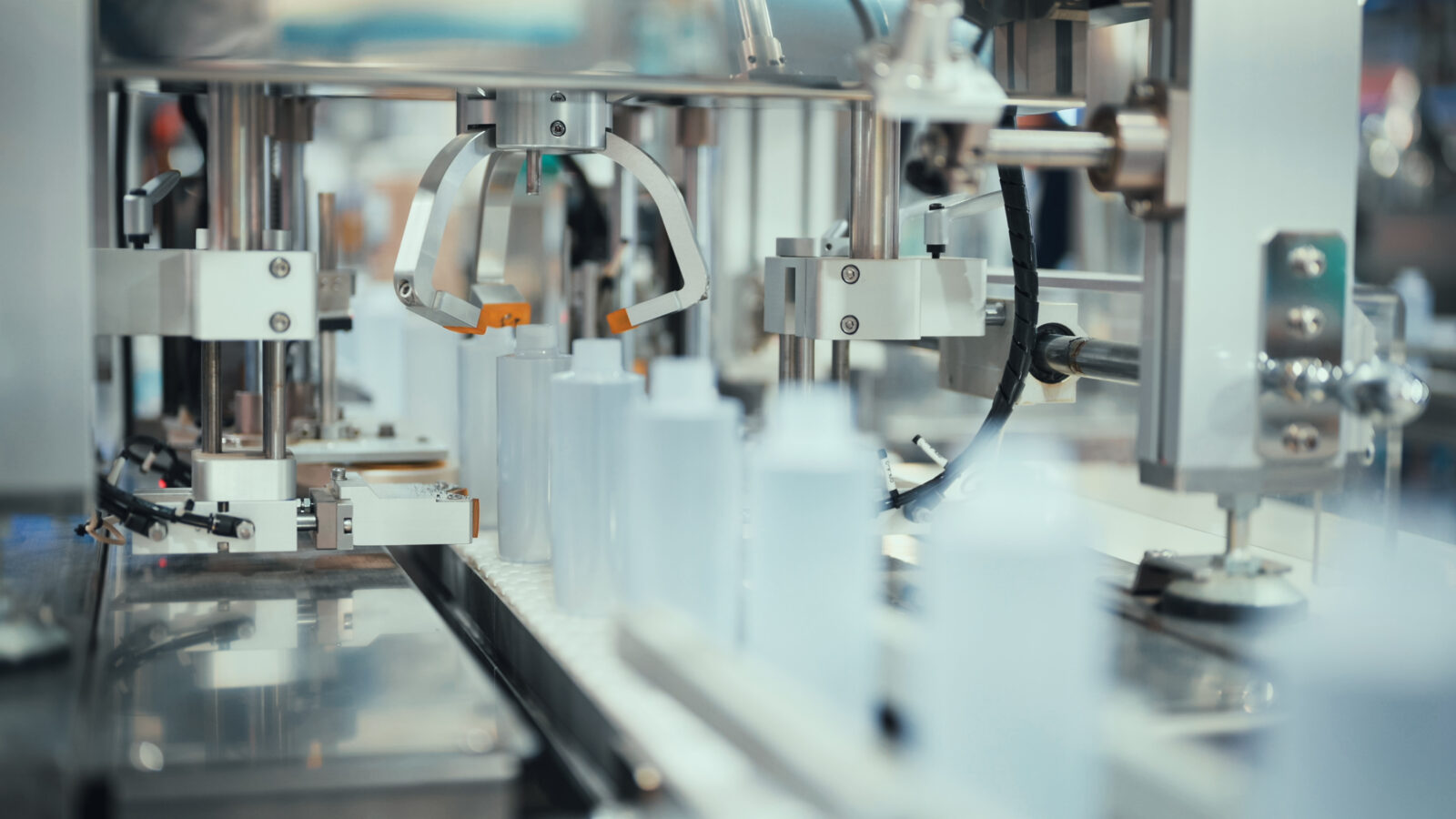 An automated robotic claw machine capping white plastic bottles on a conveyor belt in a manufacturing facility.