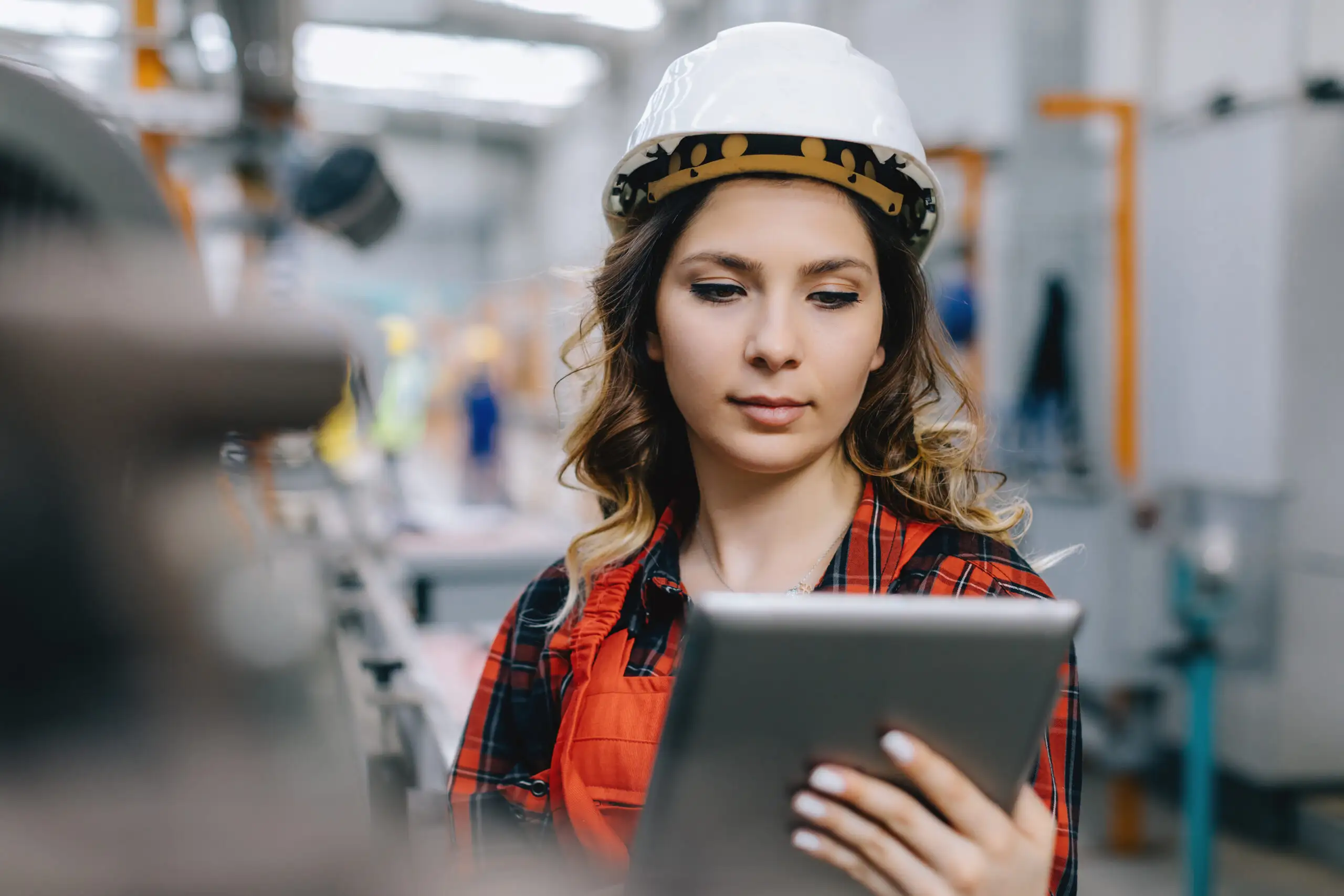 A factory worker wearing a white hard hat and red plaid shirt operating a digital tablet on an industrial plant floor.