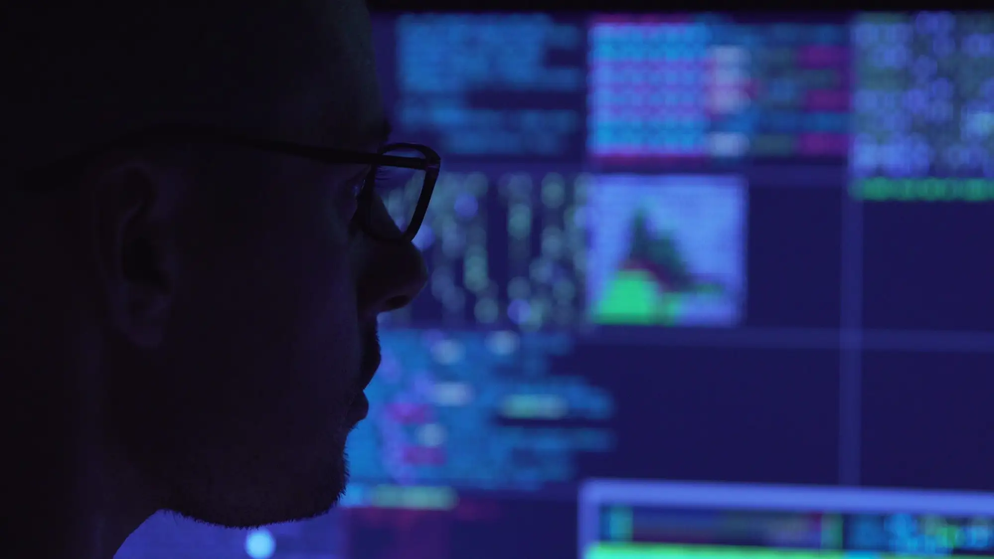 A close-up profile of a person wearing glasses in low lighting, looking attentively at a monitor displaying glowing computer code and data screens.