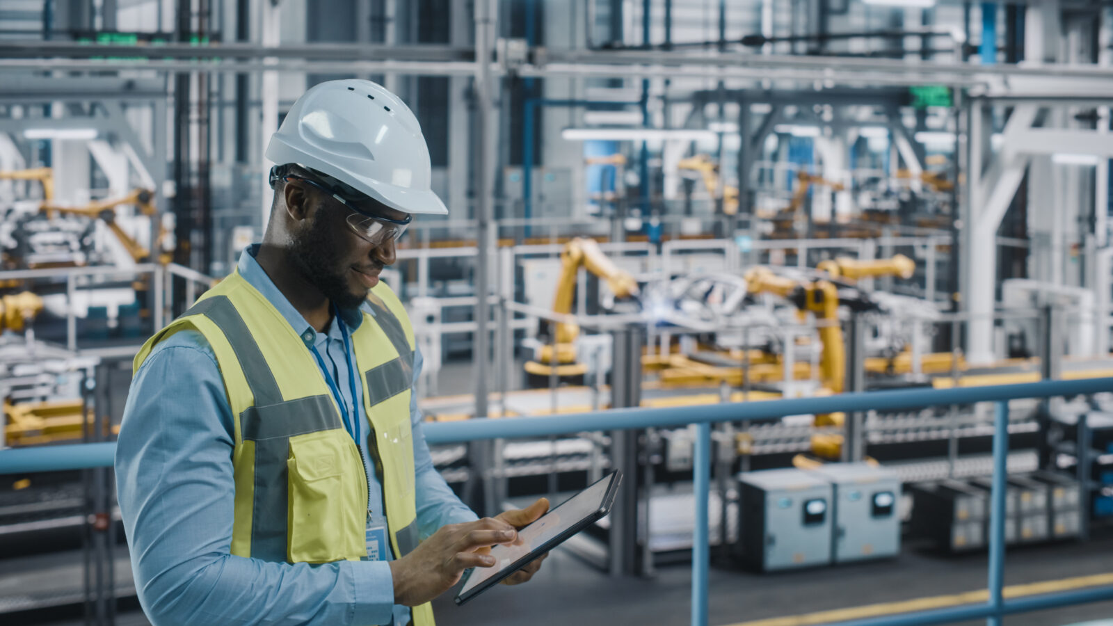 A male engineer in a white hard hat, safety glasses, and reflective vest standing in a modern automated factory while using a digital tablet.