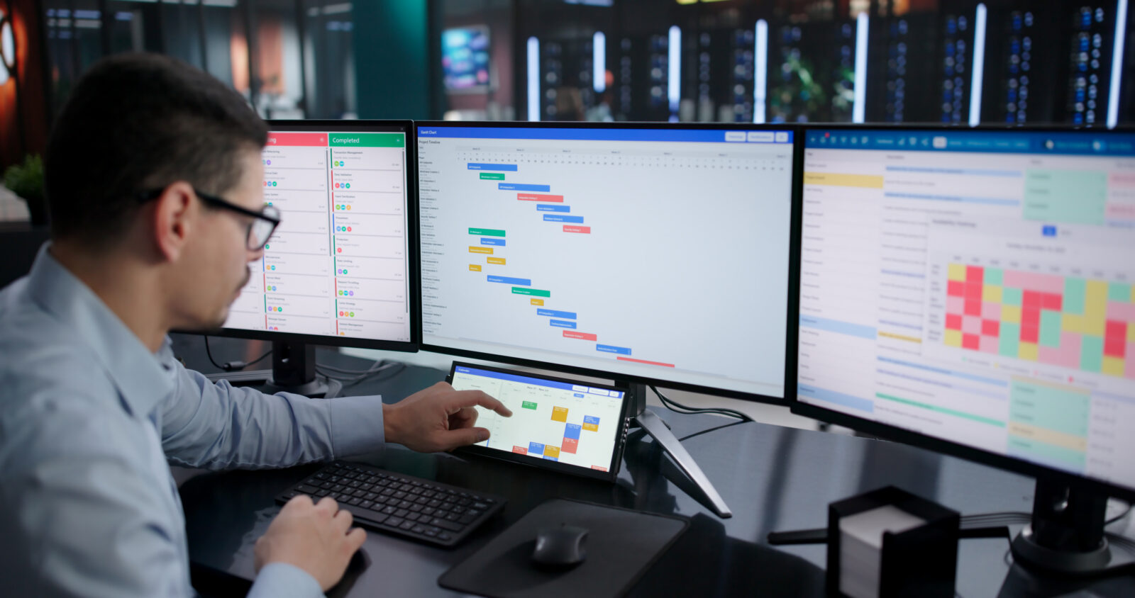 A professional working at a multi-monitor workstation with project management charts, a Gantt timeline, and a small touchscreen calendar display.