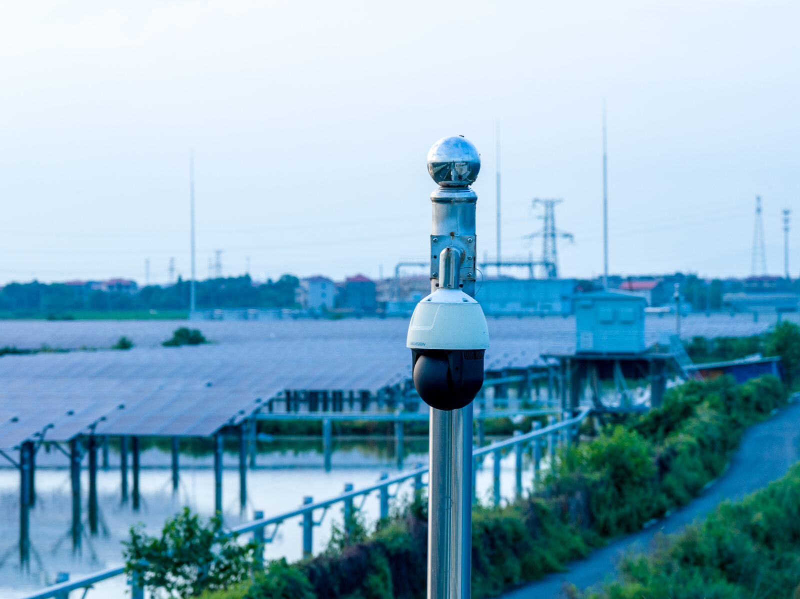 A outdoor surveillance security dome camera mounted on a metal pole overlooking a solar panel farm and industrial facility.