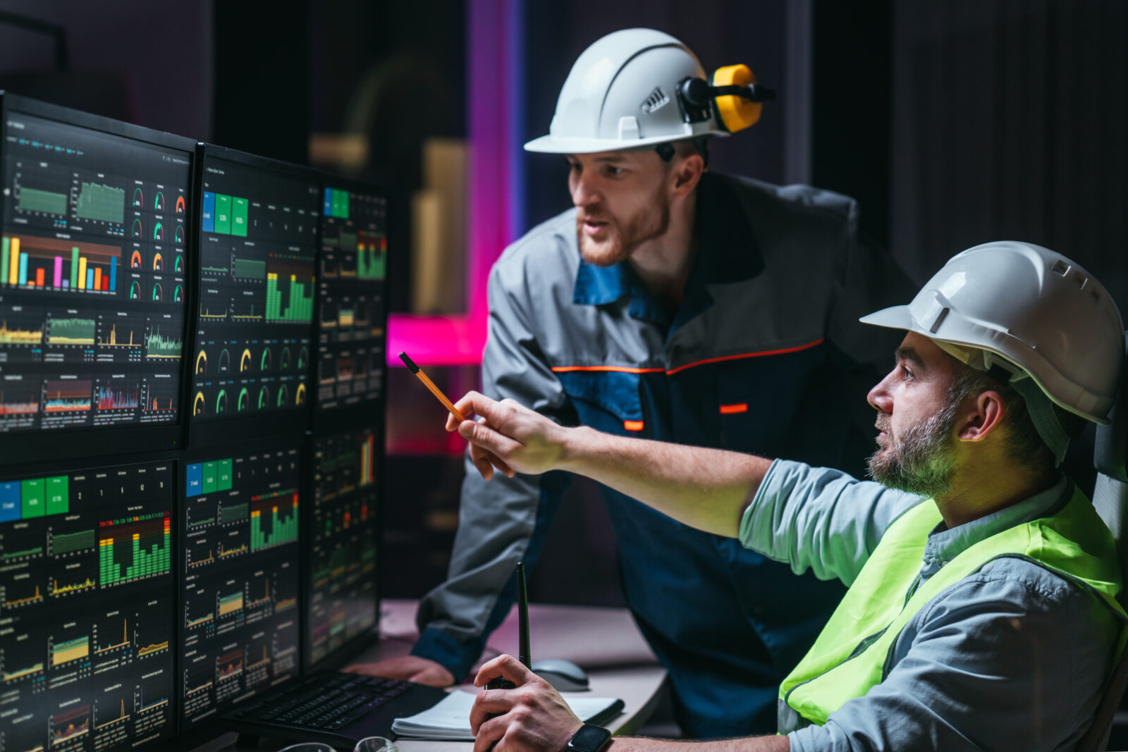 Two engineers wearing hard hats in a control room, pointing at a wall of computer screens displaying colorful data dashboards and graphs.